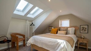 A bedroom with a sloped ceiling, skylights, a bed with white bedding, a wooden chair, a bedside table, and a small shelf. - Church Road Retreats - Coral Cottage (Kessingland)