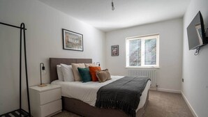 A bedroom with a bed, bedside table, lamp, window, and wall-mounted TV. - Church Road Retreats - The Cove (Kessingland)
