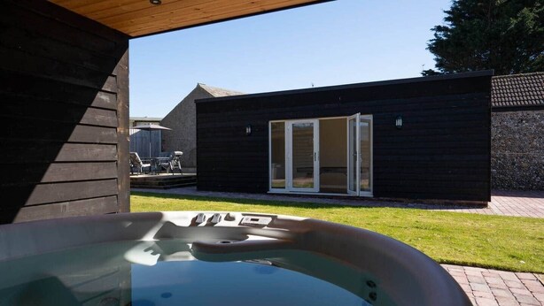 A hot tub in a backyard with a wooden deck, a patio with outdoor furniture, and a building with a sliding glass door. - Church Road Retreats - The Cove (Kessingland)