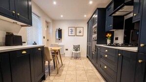 A modern kitchen with dark cabinetry, a dining area with a table and chairs, and a small white chair. - Church Road Retreats - The Cove (Kessingland)