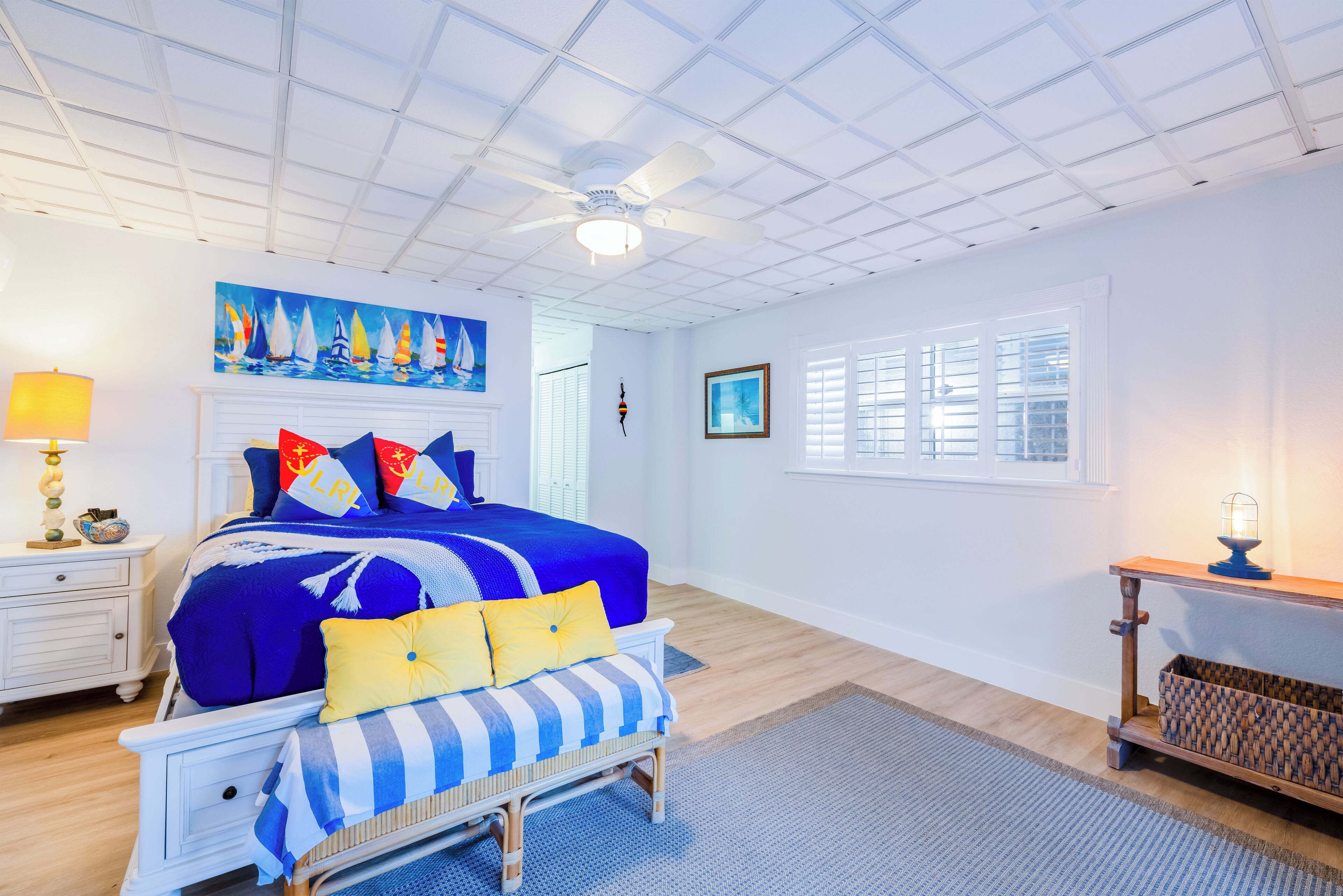 Quarto com cama grande, ventoinha no teto, janela com persianas, mesa de cabeceira com abajur e parede com pintura.