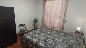 Een slaapkamer met een bed, een kledingkast, een stoel, een ventilator en een raam met gordijnen.