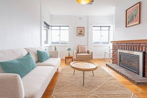Een woonkamer met een bank, fauteuil, salontafel, open haard en grote ramen.