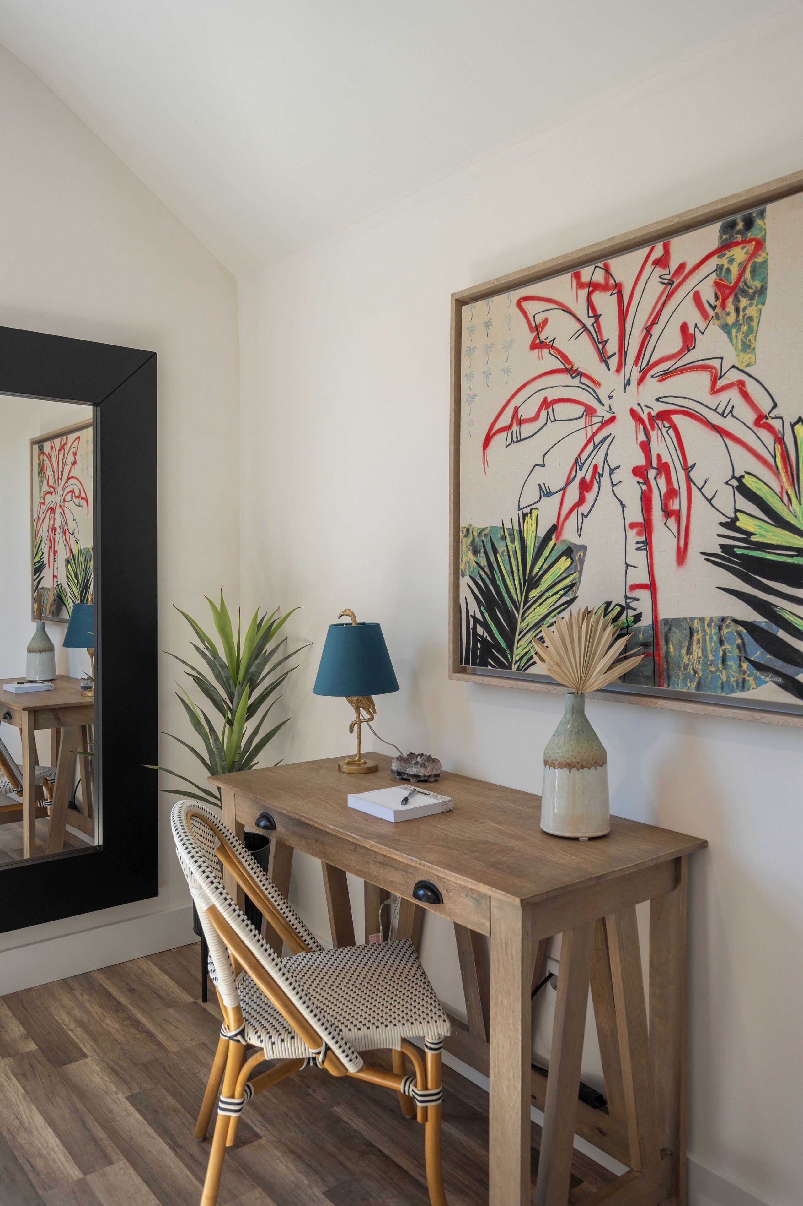A wooden desk with a chair, a lamp, and a vase. A large painting of plants is on the wall.