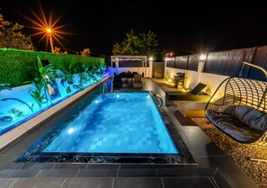 Ein Schwimmbecken mit geometrischem Fliesenmuster, umgeben von einer angelegten Grünfläche mit Pflanzen und einer Pergola.
