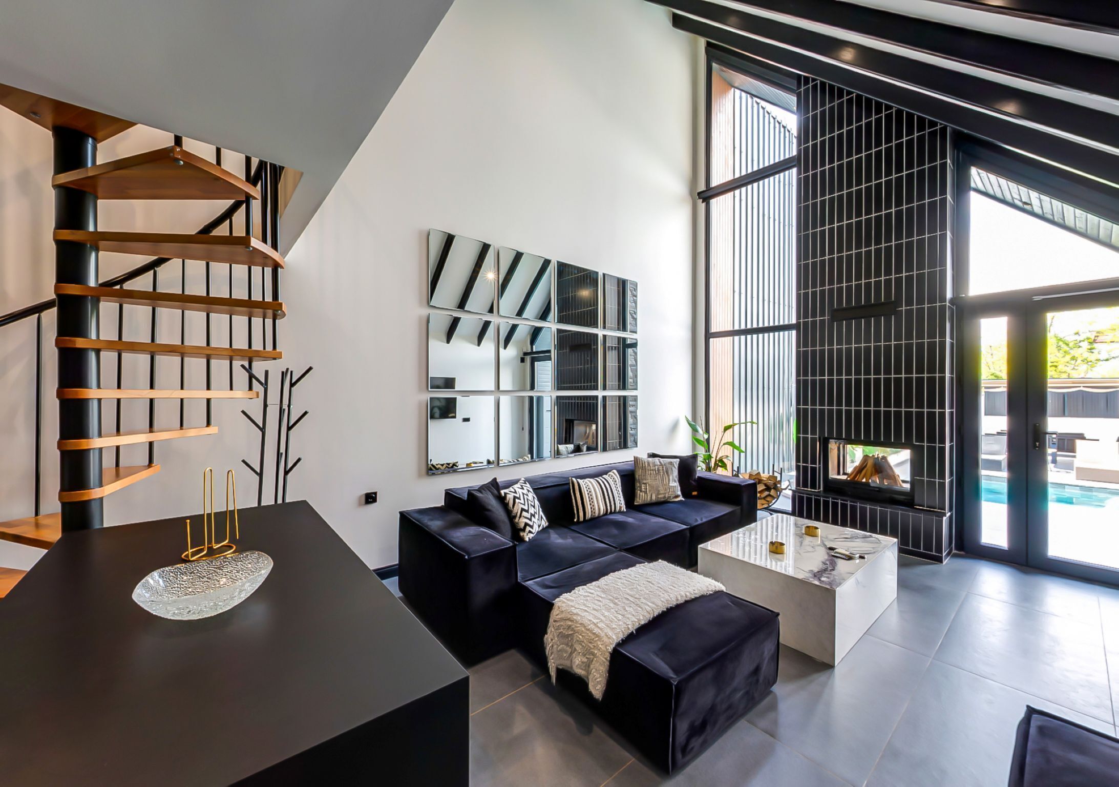 A modern living room with a black sectional sofa, a glass coffee table, and a spiral staircase.