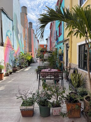 Une ruelle étroite bordée d’immeubles aux couleurs vives, de plantes en pot et d’un banc avec une table.