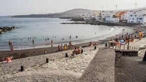 Een strand met mensen die zwemmen en zonnebaden, een stenen pad en windturbines op de achtergrond.