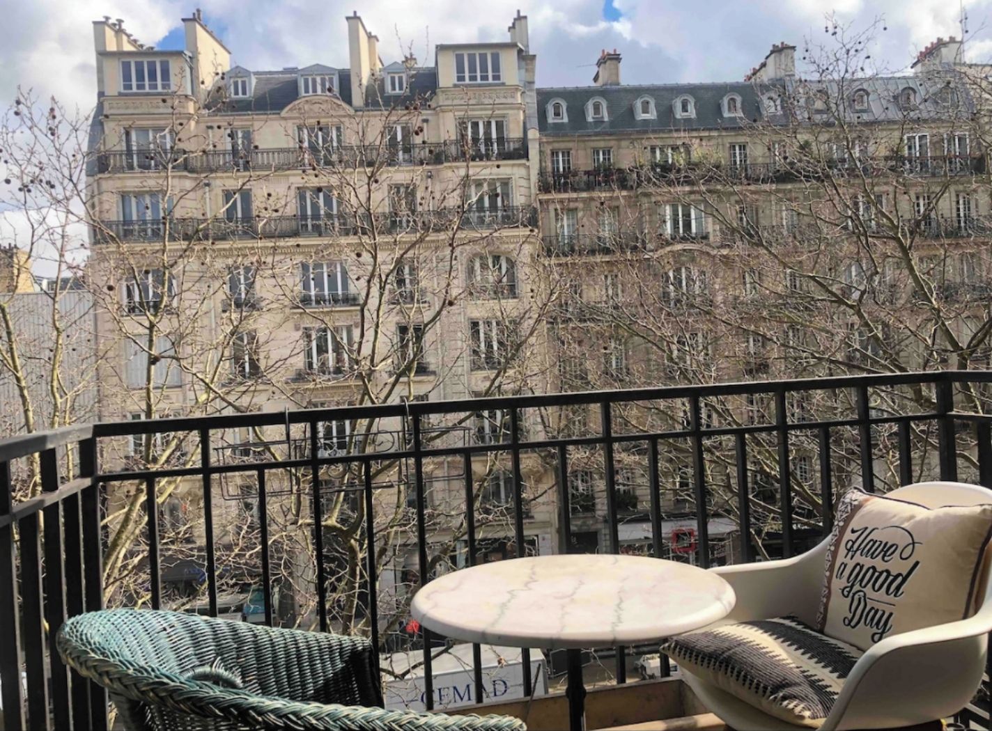 Un balcon donnant sur les immeubles parisiens, une chaise en osier, une petite table ronde et un coussin orné d’un message.
