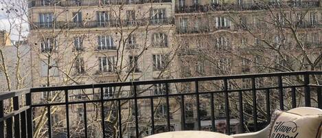 Un balcon donnant sur les immeubles parisiens, une chaise en osier, une petite table ronde et un coussin orné d’un message.