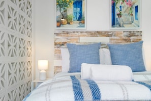 A neatly arranged bedroom with a white bed, blue pillows, and a white nightstand with a lamp. - Studio on North (Cape Town)