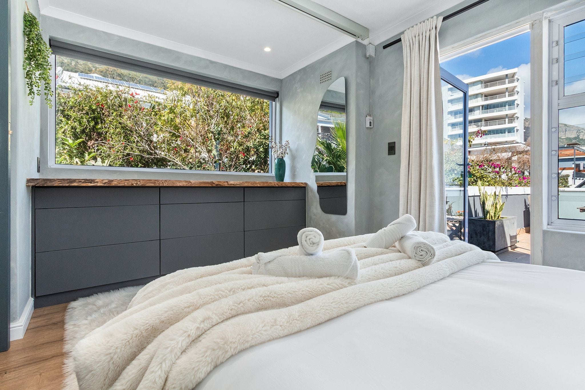 A modern bedroom with a large bed, a view of greenery outside, and a balcony with plants.