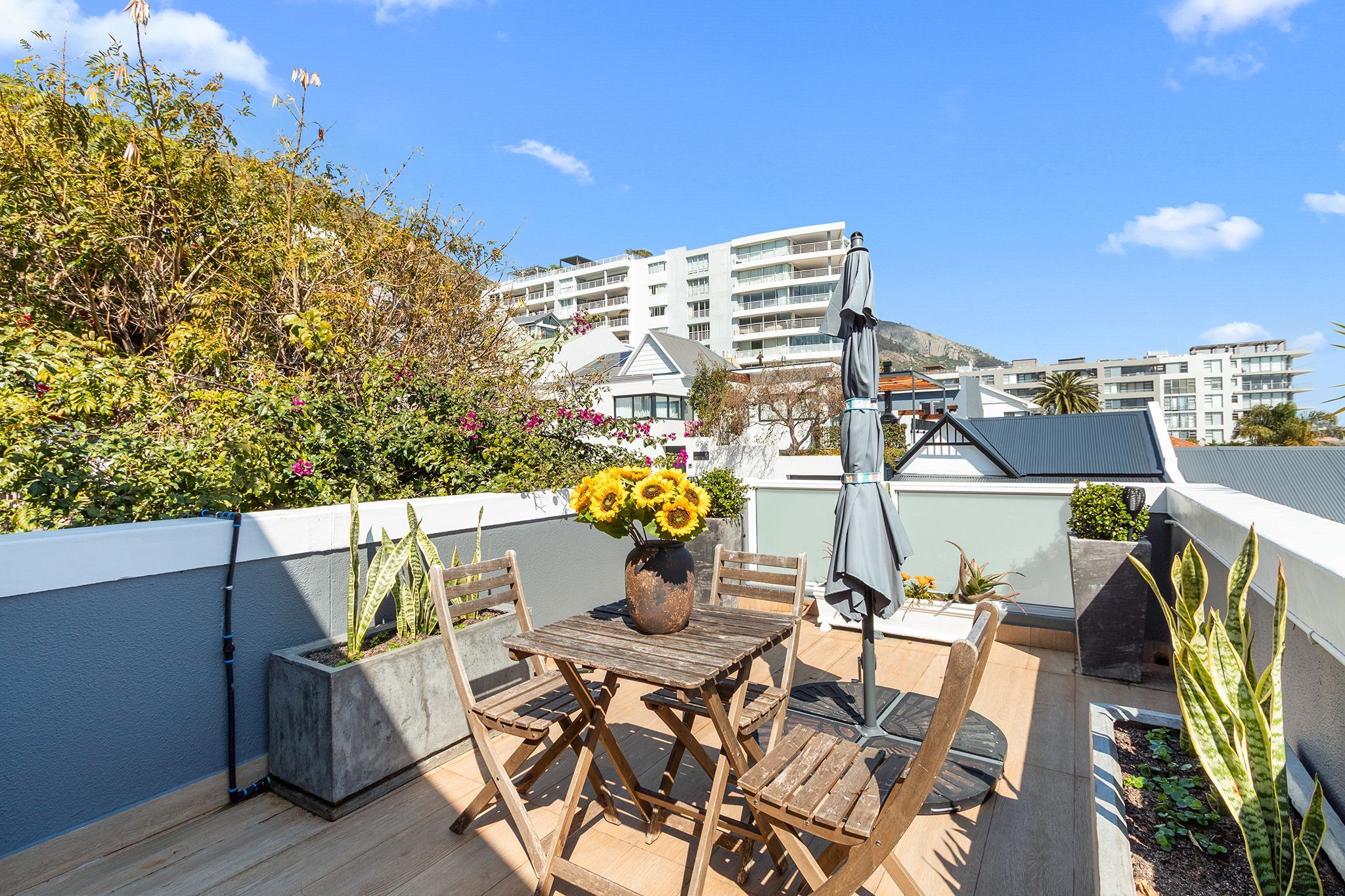 Une terrasse sur le toit avec une table et des chaises en bois, un vase rempli de tournesols et, en arrière-plan, la vue d’un bâtiment moderne.