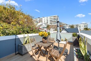 A rooftop terrace with a wooden table and chairs, a vase of sunflowers, and a view of a modern building in the background. - Selbourne Five (Cape Town)