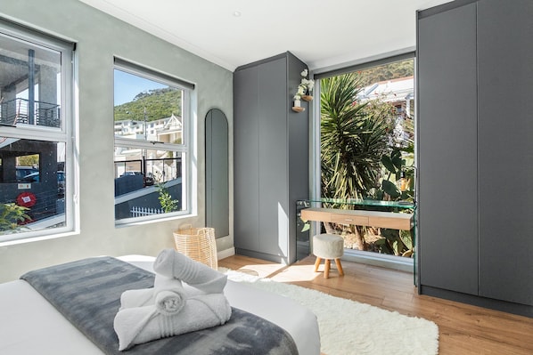 A modern bedroom with a large window, a bed with a white and gray blanket, a wooden bench, and a small table with a basket. - Selbourne Five (Cape Town)