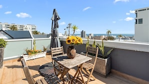 Eine Dachterrasse mit Holztisch und -stühlen, einer Vase mit Sonnenblumen und Topfpflanzen.