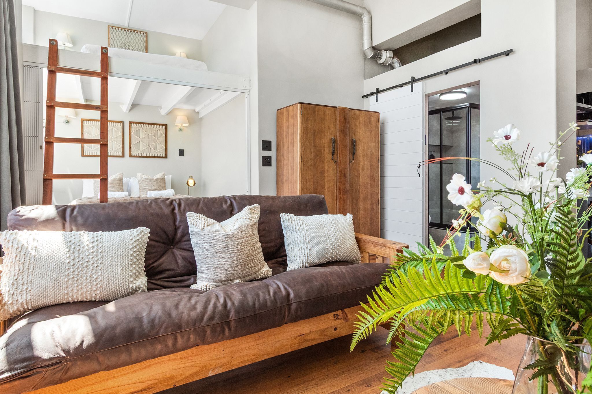 A modern living room with a brown leather sofa, white textured cushions, a wooden ladder, a large wooden cabinet, and a green fern plant.