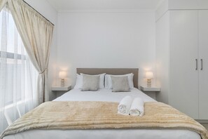 Ein ordentlich bezogenes Bett mit einer flauschigen, beige farbenen Decke, zwei Nachttischlampen und einem Paar zusammengerollter Handtücher.
