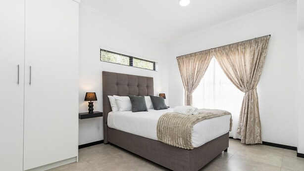 A modern bedroom with a large bed, a nightstand, a wall-mounted light, and a window with curtains. - Mandalay Hideaway (Johannesburg)