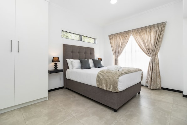 A modern bedroom with a large bed, a nightstand, a wall-mounted light, and a window with curtains.