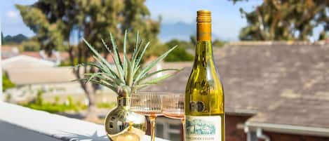 A bottle of wine, a glass, and a plant on a balcony with a view of trees and buildings.