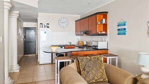 A compact kitchen with a refrigerator, microwave, and wooden cabinets. A clock is mounted on the wall, and a sign with directional arrows is visible. - Bali Hai 103 (Westbrook)