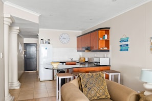 A compact kitchen with a refrigerator, microwave, and wooden cabinets. A clock is mounted on the wall, and a sign with directional arrows is visible. - Bali Hai 103 (Westbrook)