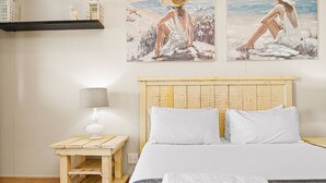 A bedroom with a wooden headboard, white bedding, a small wooden nightstand with a lamp, and two beach-themed paintings on the wall. - Bali Hai 103 (Westbrook)