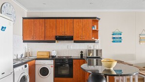 A compact kitchen with wooden cabinets, a built-in oven, a washing machine, and a small dining area with a table and stools. - Bali Hai 103 (Westbrook)