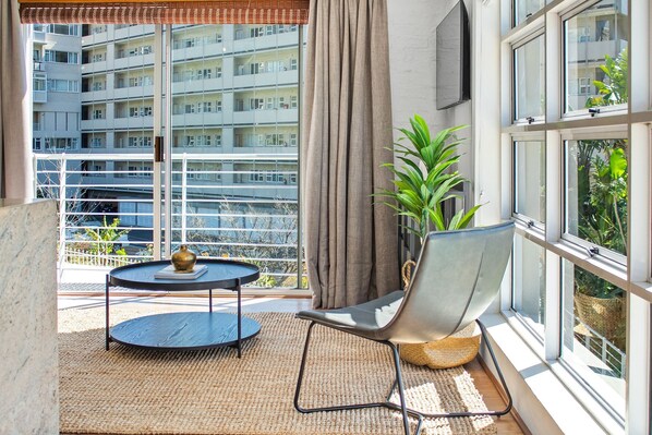 Un balcón con vista a un edificio alto, una mesa redonda con un florero, una silla y una planta en maceta.