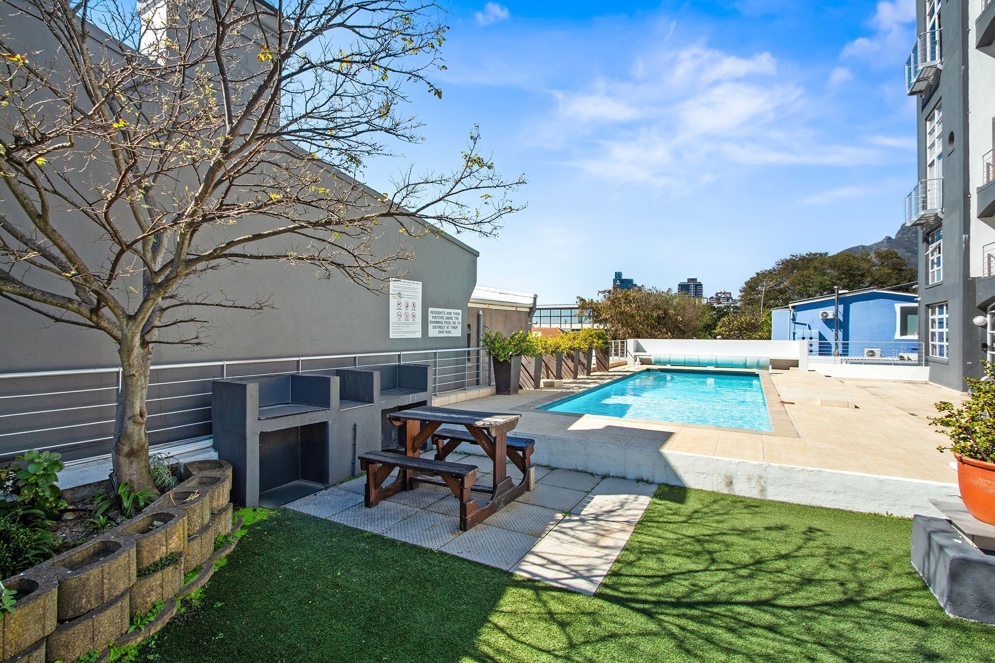 A modern outdoor pool area with a wooden table and benches, surrounded by a well-maintained lawn and a tree.