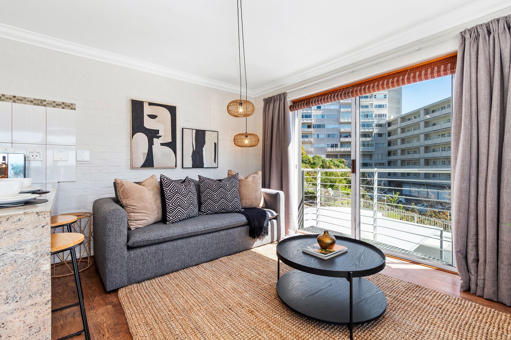 Una sala moderna con un sofá gris, cojines blancos y negros, una mesa de centro redonda y vistas de un paisaje urbano a través de amplios ventanales.