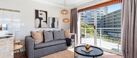 Una sala moderna con un sofá gris, cojines blancos y negros, una mesa de centro redonda y vistas de un paisaje urbano a través de amplios ventanales.