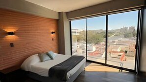 A modern bedroom with a large bed, wooden headboard, and a city view through floor-to-ceiling windows.