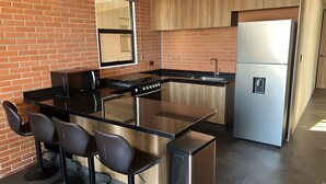 A modern kitchen with a brick wall, a stainless steel refrigerator, a black countertop, and bar stools.