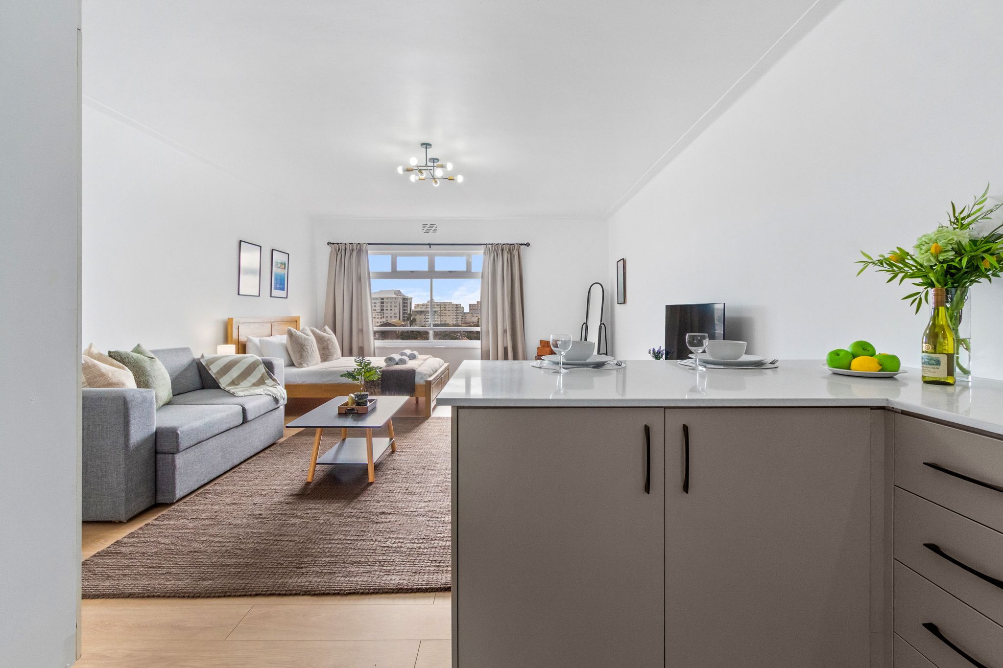 A modern living room with a grey sofa, a coffee table, and a view of the city through large windows.