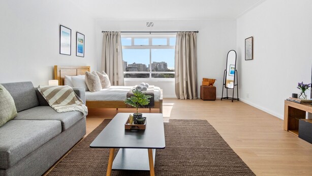 A modern living room with a sofa, a bed, a coffee table, and a view of the city through large windows. - E1 Ventnor House (Cape Town)
