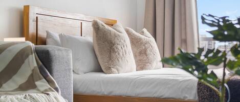 A bed with a wooden headboard, white bedding, and two fluffy pillows. There is a gray armchair with a striped throw blanket. A potted plant is visible in the foreground.