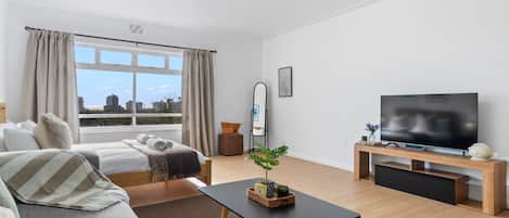 A modern hotel room with a bed, sofa, TV, and a view of the city.
