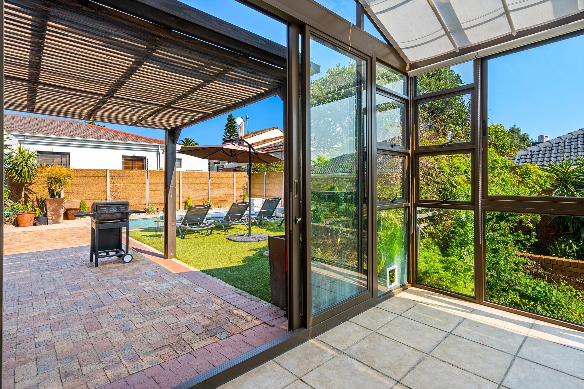 Un espace extérieur abrité, équipé d’un barbecue, de chaises longues et offrant une vue sur un jardin avec une piscine.
