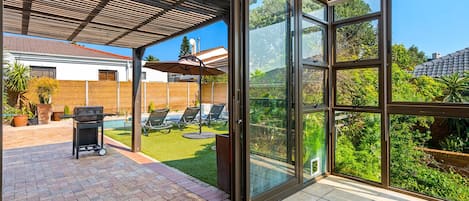 Un espace extérieur abrité, équipé d’un barbecue, de chaises longues et offrant une vue sur un jardin avec une piscine.