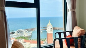 A hotel room with a large window offering a view of the sea and a bridge. There is a chair with a patterned cushion and a bed with a white cover. - Atermis Regency Hillside Phu Quoc - Sea & Fireworks View (Phu Quoc)