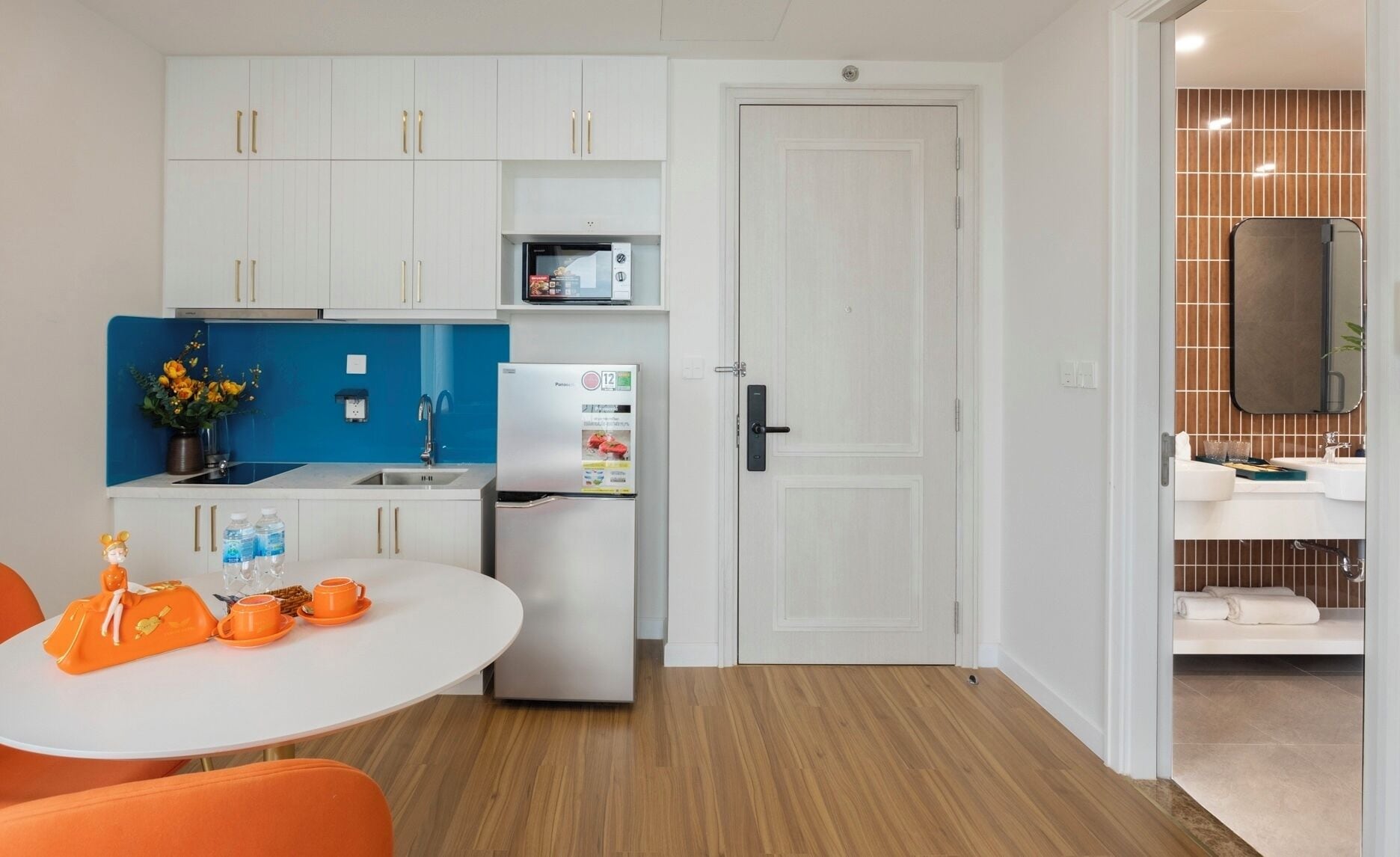 Uma cozinha compacta com armários brancos, geladeira, micro-ondas e pia. Há uma área de refeições com uma mesa branca e cadeiras laranja, e um pequeno objeto decorativo sobre a mesa.