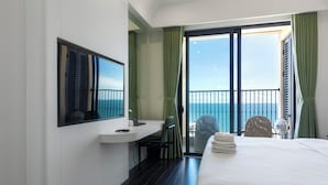 A hotel room with a bed, a desk, a chair, a balcony with a view of the sea, and a large window with curtains. - Atermis Regency Hillside Phu Quoc - Sea & Fireworks View (Phu Quoc)