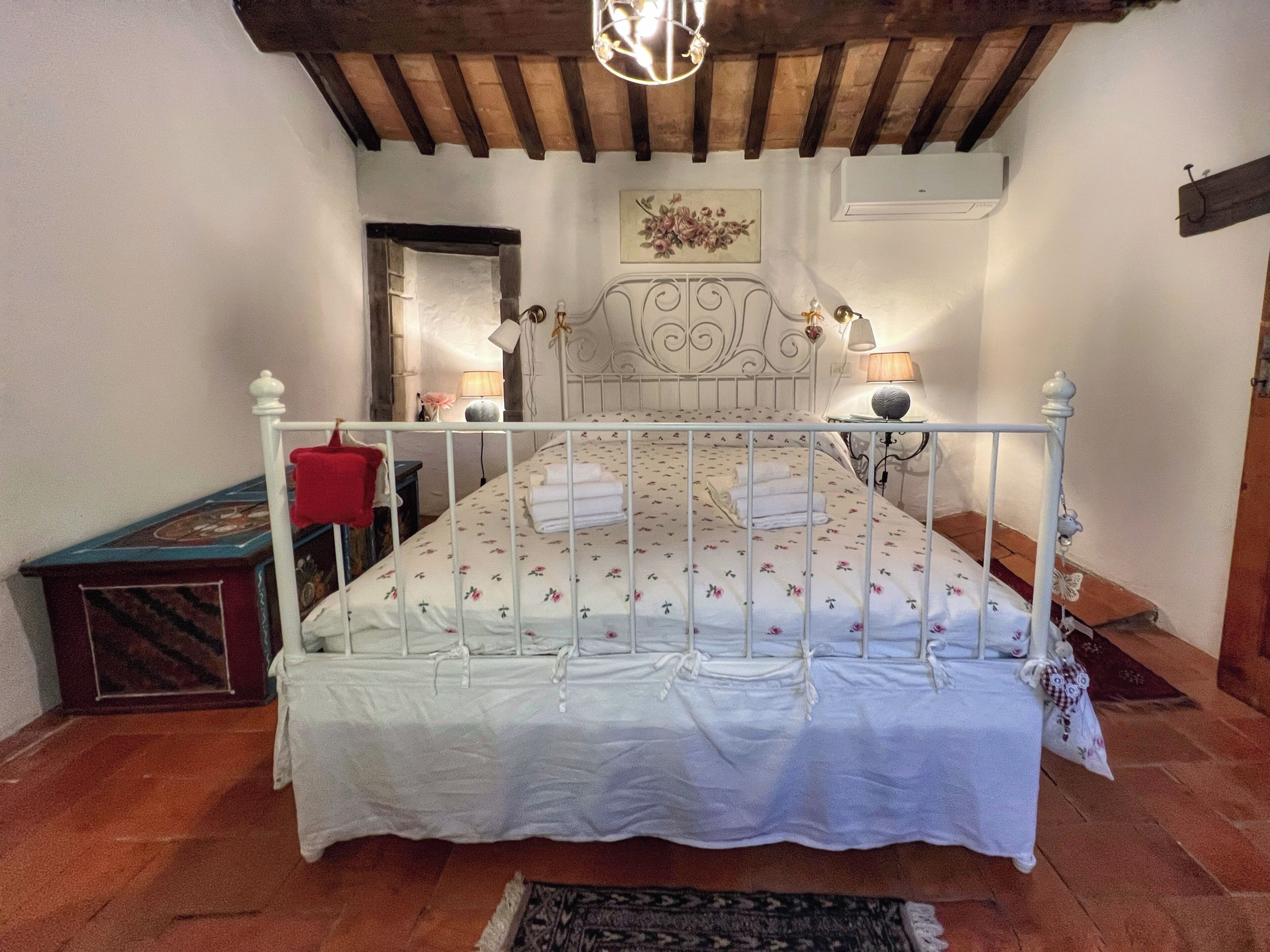 Ein Schlafzimmer mit weißem Metallbettgestell, floralem Bettzeug, Holzboden, einem roten Sessel und einem kleinen Tisch mit Lampe.