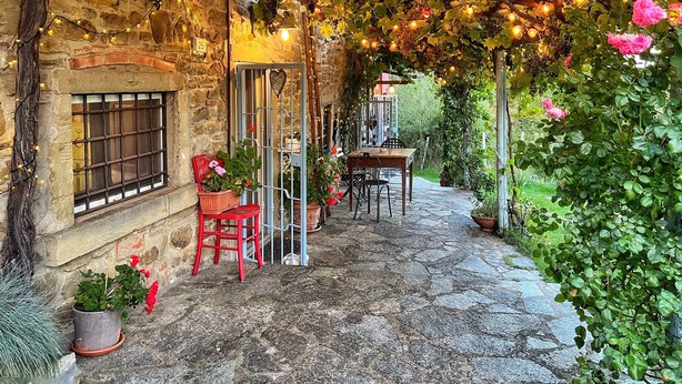 A stone building with a rustic stone floor, a wooden table and chairs under a vine-covered pergola with string lights, and potted plants. - La Fonte - Two Bedroom House, Sleeps 4 (Subbiano)
