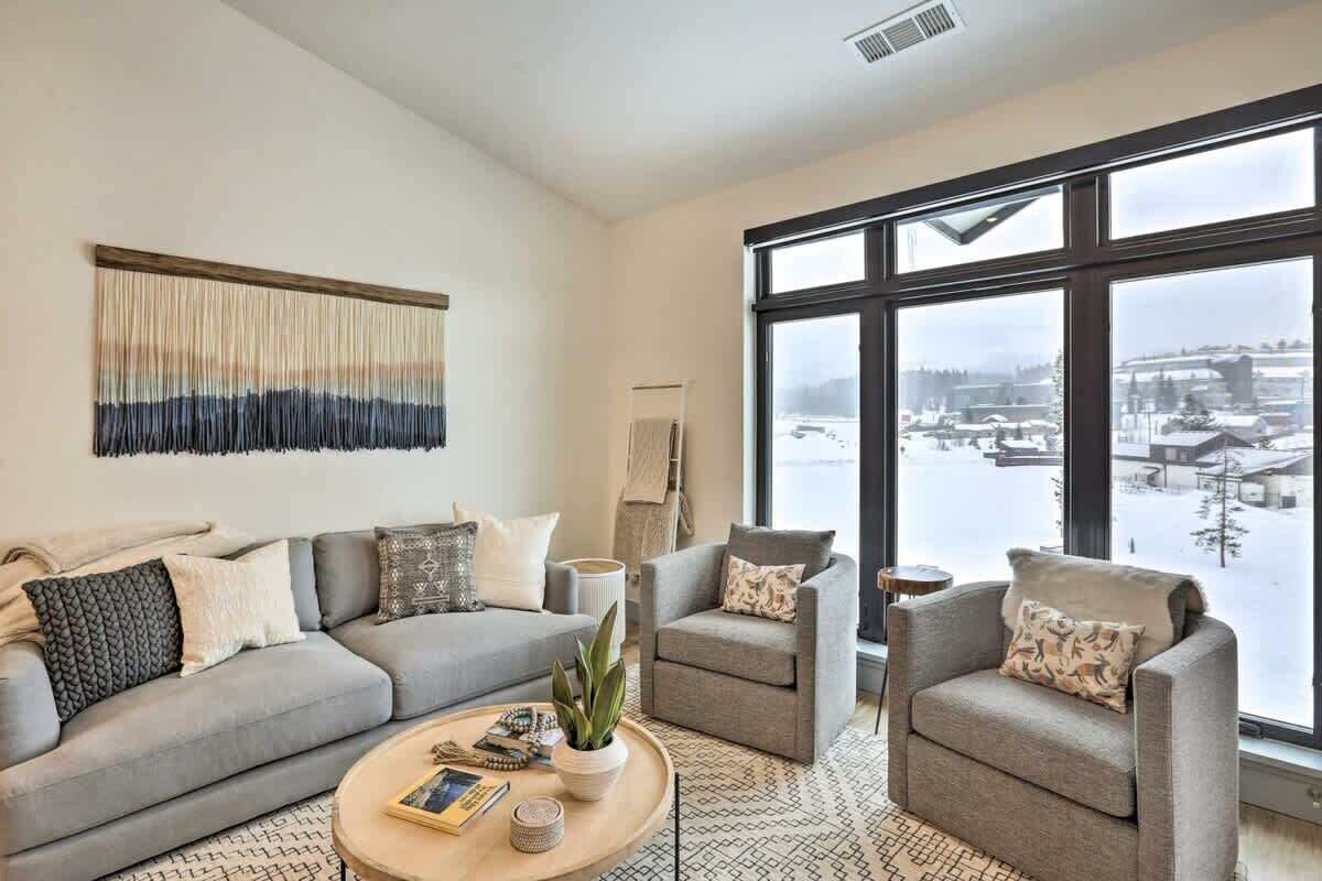Una sala de estar moderna con un sofá gris, dos sillones, una mesa de centro y un ventanal con vistas a un paisaje nevado.