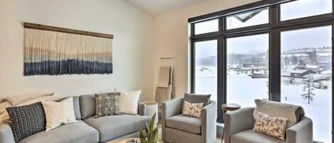 Una sala de estar moderna con un sofá gris, dos sillones, una mesa de centro y un ventanal con vistas a un paisaje nevado.