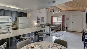 Una cocina moderna con una isla central, un área de comedor con mesa redonda y una sala de estar con sofá y ventilador de techo.