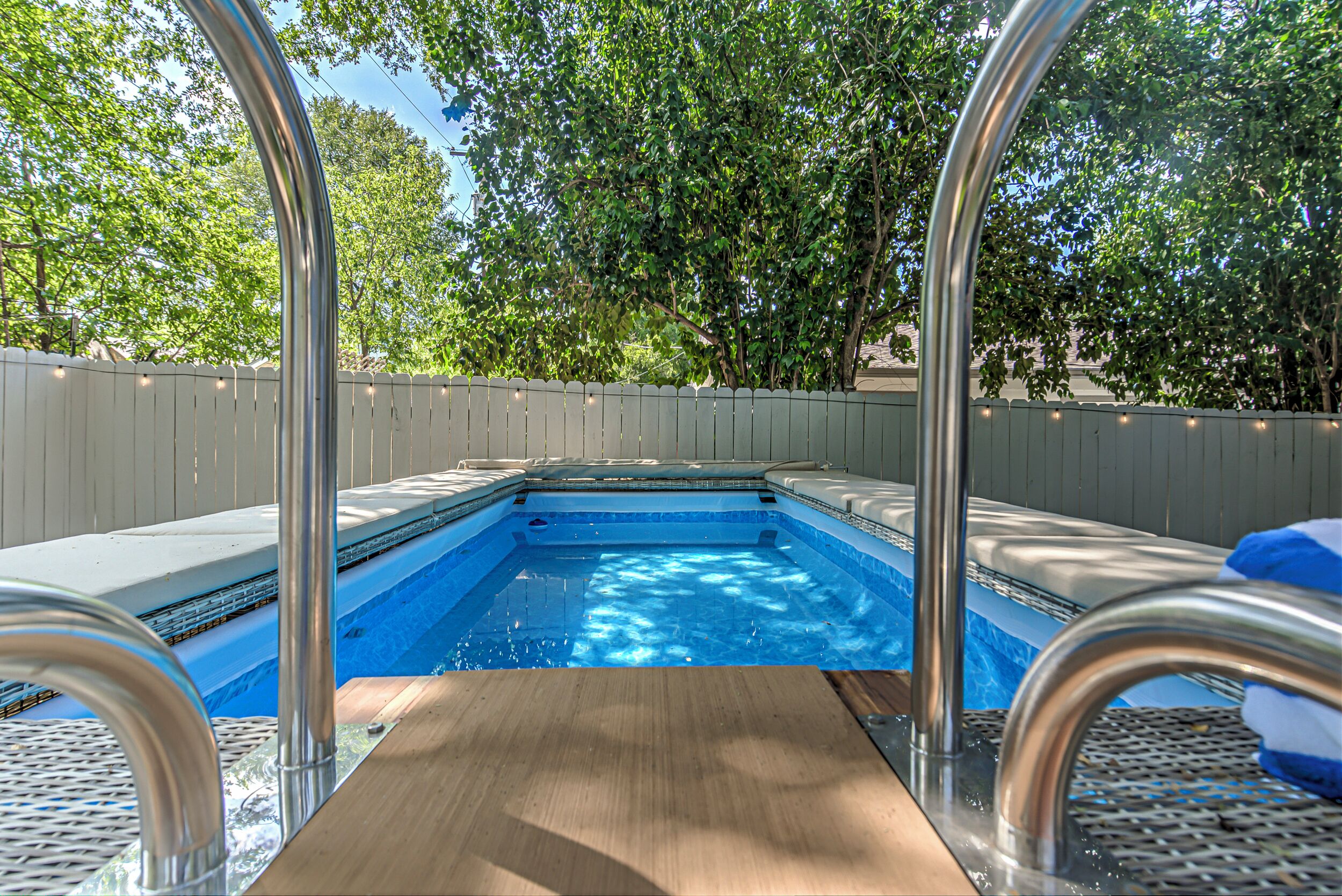 A swimming pool with a wooden deck and metal railings, surrounded by a white fence and trees.